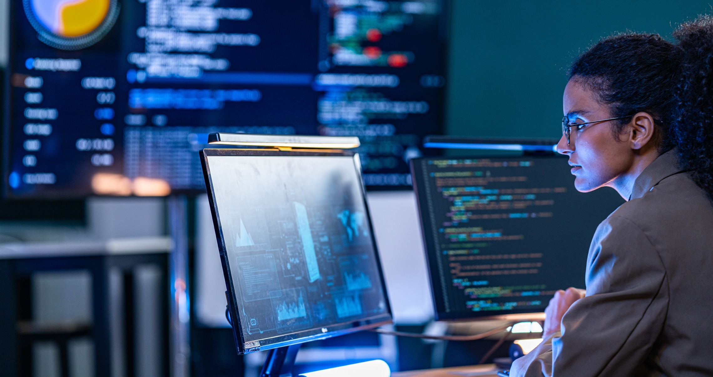 A woman wearing glasses works at a computer workstation surrounded by monitors displaying code, data visualizations, and dashboards in blue lighting.